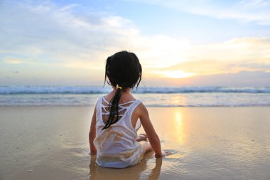 Asyalı kız görünümünü geri oturma ve oynamaya gün batımında sahilde kum.