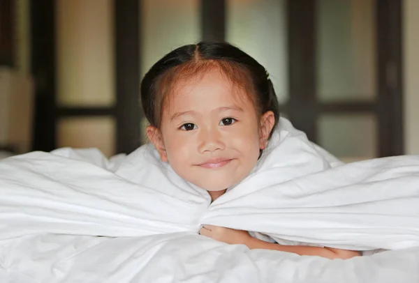Happy little asian girl lying under blanket on the bed