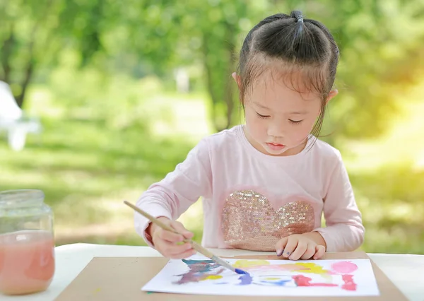 Child painting, little girl having fun to paint on paper in the green garden.