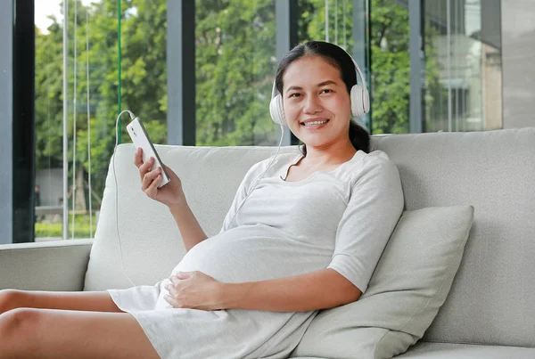 Pregnant woman is sitting and relax on sofa listening music in headphones.