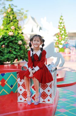 Kırmızı elbiseli mutlu Asyalı küçük kız arka planda Noel ağacına karşı büyük bir hediye kutusunda oturuyor..
