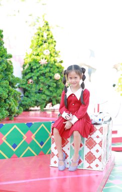 Kırmızı elbiseli mutlu Asyalı küçük kız arka planda Noel ağacına karşı büyük bir hediye kutusunda oturuyor..