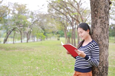 Kitap açık parkta ağaç gövdesi karşı yalın ayakta Portre genç Asyalı kadın.