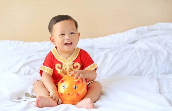 自宅でベッドに座って貯金箱と伝統的な中国のドレスで幸せな小さなアジアの赤ちゃんの男の子 子供の貯金の概念 — ストック写真