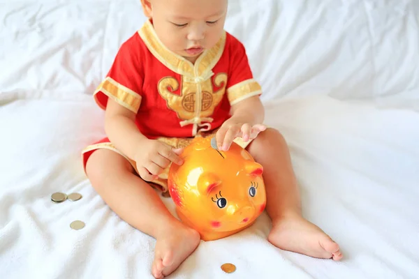 关闭穿着中国传统服装的亚洲小男孩 把一些硬币放进坐在家里床上的存钱罐里 孩子省钱的概念 专注于存钱罐 — 图库照片