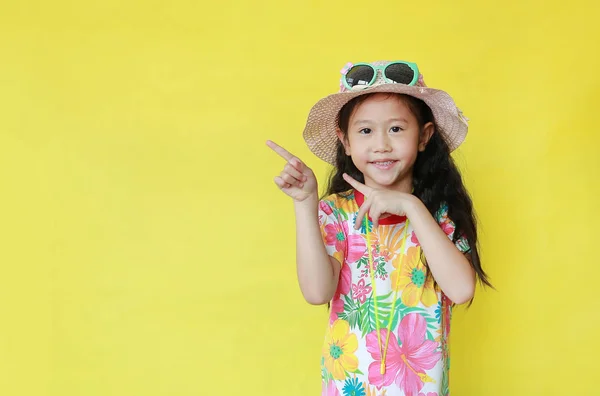 コピースペースと黄色の背景に隔離された陽気な女の子を指しています 花模様の夏のドレスとサングラスで帽子の子供 休日と夏のファッションコンセプト — ストック写真