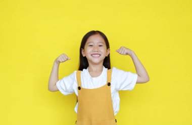 Güçlü Asyalı küçük kız ellerini kaldırıp gülümsüyor. Sarı arka planda izole edilmiş kot pantolonlu mutlu çocuk portresi..