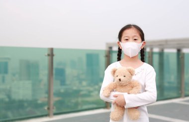 Coronavirus covid-19 ve kirlilik koruma konsepti. Asyalı küçük kız, Bangkok 'ta Corona virüsü ve hava kirliliğine karşı maske takan oyuncak ayıya sarılıyor. Tayland.