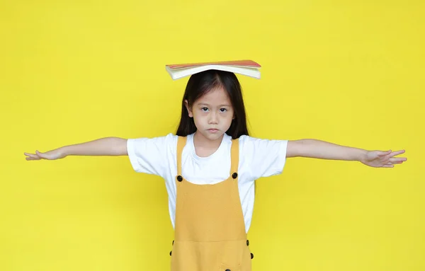黄色の背景に隔離された頭の上に本を持つ不幸なアジアの子供の女の子 罰を受けた女子高生の肖像画が腕を広げカメラを見て — ストック写真