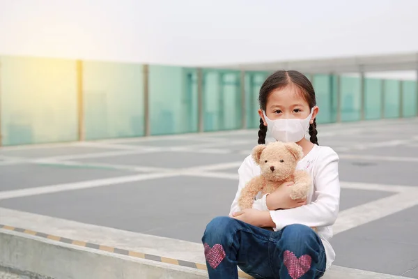 Coronavirus covid-19 ve kirlilik koruma konsepti. Asyalı küçük kız, Bangkok 'ta Corona virüsü ve hava kirliliğine karşı maske takan oyuncak ayıya sarılıyor. Tayland.