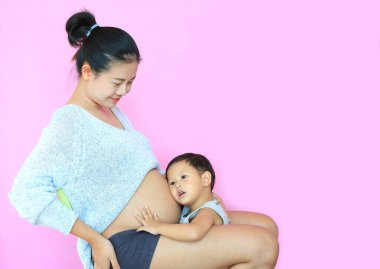 Pembe arka planda izole edilmiş hamile bir annenin karnındaki bebeği dinleyen Asyalı bebek. Hamilelik ve yeni yaşam konsepti