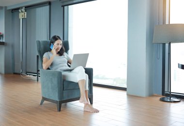 Akıllı telefon ve dizüstü bilgisayar kullanan genç ve güzel Asyalı kadın evde çalışıyor ve koltukta dinleniyor..