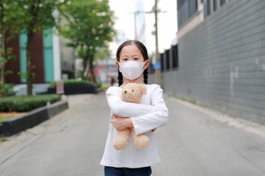 Corona virüsünü ve hava kirliliğini korumak için maske takan Asyalı küçük kız portresi dışarıda duran oyuncak ayıyla birlikte pm2.5 tozu. Coronavirus covid-19 ve kirlilik koruma kavramı