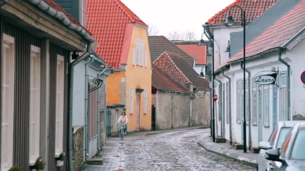 Kuressaare, Ösel, Estland. Kvinna cykling på Kohtu Street i molnigt vinterdag. Gamla traditionella hus på smala gator — Stockvideo