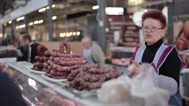 Литва, Вильнюс. Продавщица на рынке Галле продает колбасу и другие мясные продукты. Продавец обслуживает покупателя на местном продовольственном рынке . — стоковое видео
