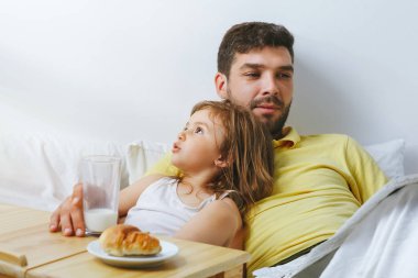 Baba ve kızı yatakta kahvaltı yapıyor. Babam gazete okur.