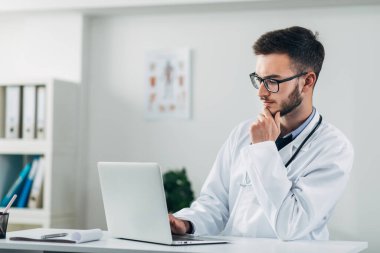 Genç doktor ofisinde dizüstü bilgisayarla çalışıyor.