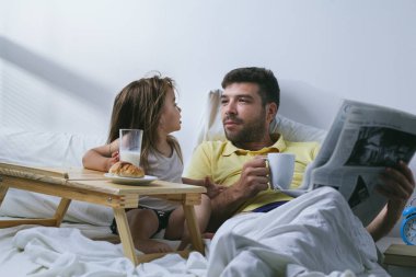Baba ve kızı sabahları yatakta kahvaltı yapıyorlar. Babam gazete okuyor.