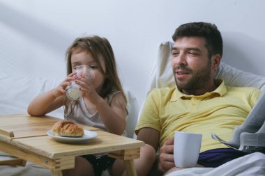 Baba ve kızı sabahları yatakta kahvaltı yapıyorlar. Babam gazete okuyor.