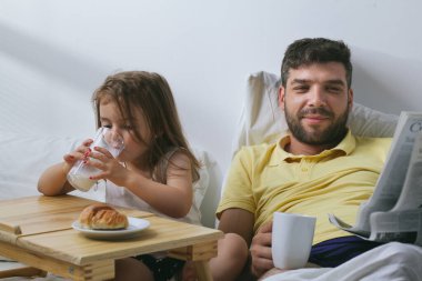 Baba ve kızı sabahları yatakta kahvaltı yapıyorlar. Babam gazete okuyor.