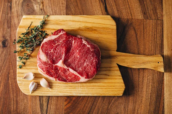 Overhead shot of ribeye steak resting on a wooden board in the kitchen with fresh thyme and garlic cloves. Top shot