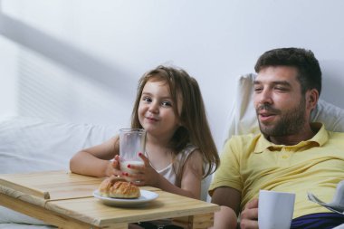 Baba ve kızı sabahları yatakta kahvaltı yapıyorlar. Babam gazete okuyor.
