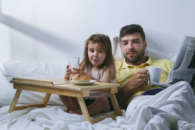 Baba ve kızı sabahları yatakta kahvaltı yapıyorlar. Babam gazete okuyor.