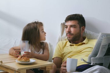 Baba ve kızı sabahları yatakta kahvaltı yapıyorlar. Babam gazete okuyor.