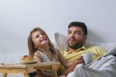 Baba ve kızı sabahları yatakta kahvaltı yapıyorlar. Babam gazete okuyor.