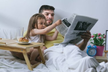Baba ve kızı sabahları yatakta kahvaltı yapıyorlar. Babam gazete okuyor.