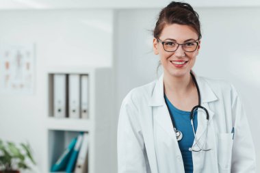 Kameraya gülümseyen genç kadın doktor portresi. Hastane ofisinde steteskobu olan dost canlısı bir doktor.