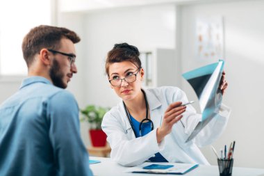 Hasta ile birlikte muayenehanesinde oturan ve röntgene bakan kadın doktorun portresi.