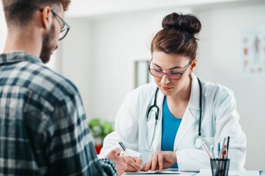 Kadın doktor, hastalığını tedavi etmesi için genç hastaya ilaç yazıyor.