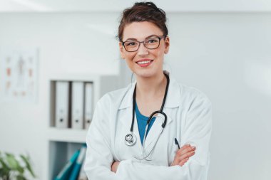 Kameraya gülümseyen genç kadın doktor portresi. Hastane ofisinde steteskobu olan dost canlısı bir doktor.