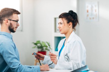Kadın doktor, steteskopla muayenehanesinde oturmuş hastasıyla konuşuyor.