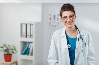 Kameraya gülümseyen genç kadın doktor portresi. Hastane ofisinde steteskobu olan dost canlısı bir doktor.