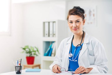 Kadın doktor stetoskopla muayenehanesinde oturuyor.