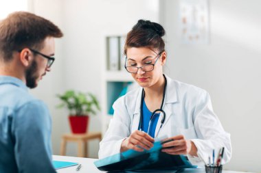 Hasta ile birlikte muayenehanesinde oturan ve röntgene bakan kadın doktorun portresi.