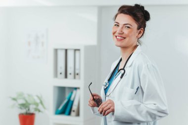 Kameraya gülümseyen genç kadın doktor portresi. Hastane ofisinde steteskobu olan dost canlısı bir doktor.