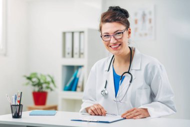 Kadın doktor stetoskopla muayenehanesinde oturuyor.