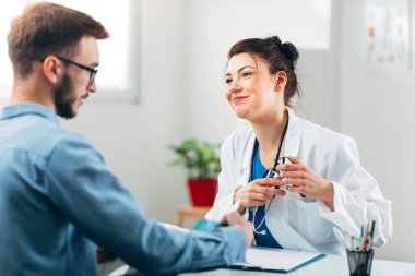 Kadın doktor, steteskopla muayenehanesinde oturmuş hastasıyla konuşuyor.