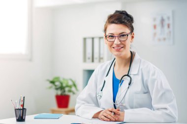 Kadın doktor stetoskopla muayenehanesinde oturuyor.