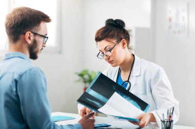 Hasta ile birlikte muayenehanesinde oturan ve röntgene bakan kadın doktorun portresi.