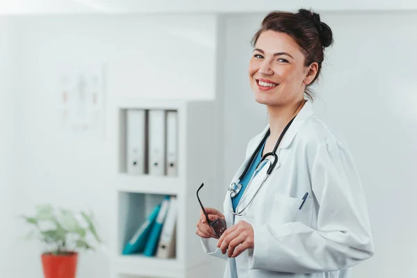 Kameraya gülümseyen genç kadın doktor portresi. Hastane ofisinde steteskobu olan dost canlısı bir doktor.