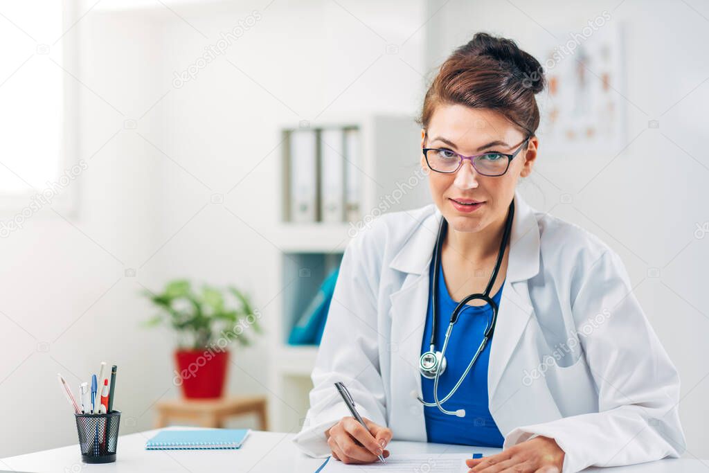 Portrait of Female Doctor sitting in her Medical Office with Stetoscope writing Prescription