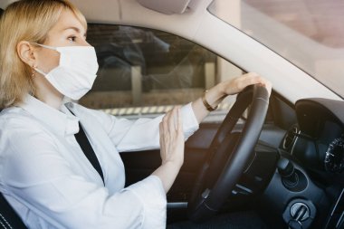 Araba kullanırken ve diğer arabalara korna çalarken koruyucu yüz maskesi takan genç bir kadın. İş kadını ofise gidiyor, Coronavirus enfeksiyonuna karşı korunuyor..