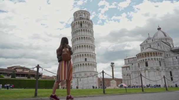 Turystka z plecakiem zbliża się do Piazza dei Miracoli, patrząc na wspaniały Leaning Tower słynny zabytek i uśmiechnięty do kamery w piękny dzień. Ogólny widok. Piza, Włochy — Wideo stockowe