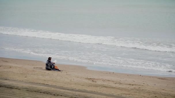 Žena sedící u moře zabalená v útulné teplé přikrývce za chladného větrného dne a čtení knihy. Na moři je mnoho vln. Boční pohled. Obecný plán — Stock video