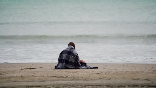 Niña sentada junto al mar envuelta en cálida manta a cuadros en un día fresco y ventoso. Hay olas en el mar. Vista trasera — Vídeos de Stock