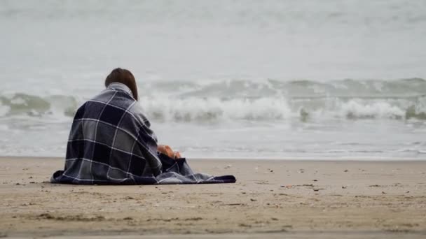 Chica morena sentada junto al mar envuelta en una cálida manta a cuadros en el ventoso y fresco día de primavera. Hay olas en el mar. Vista trasera — Vídeos de Stock